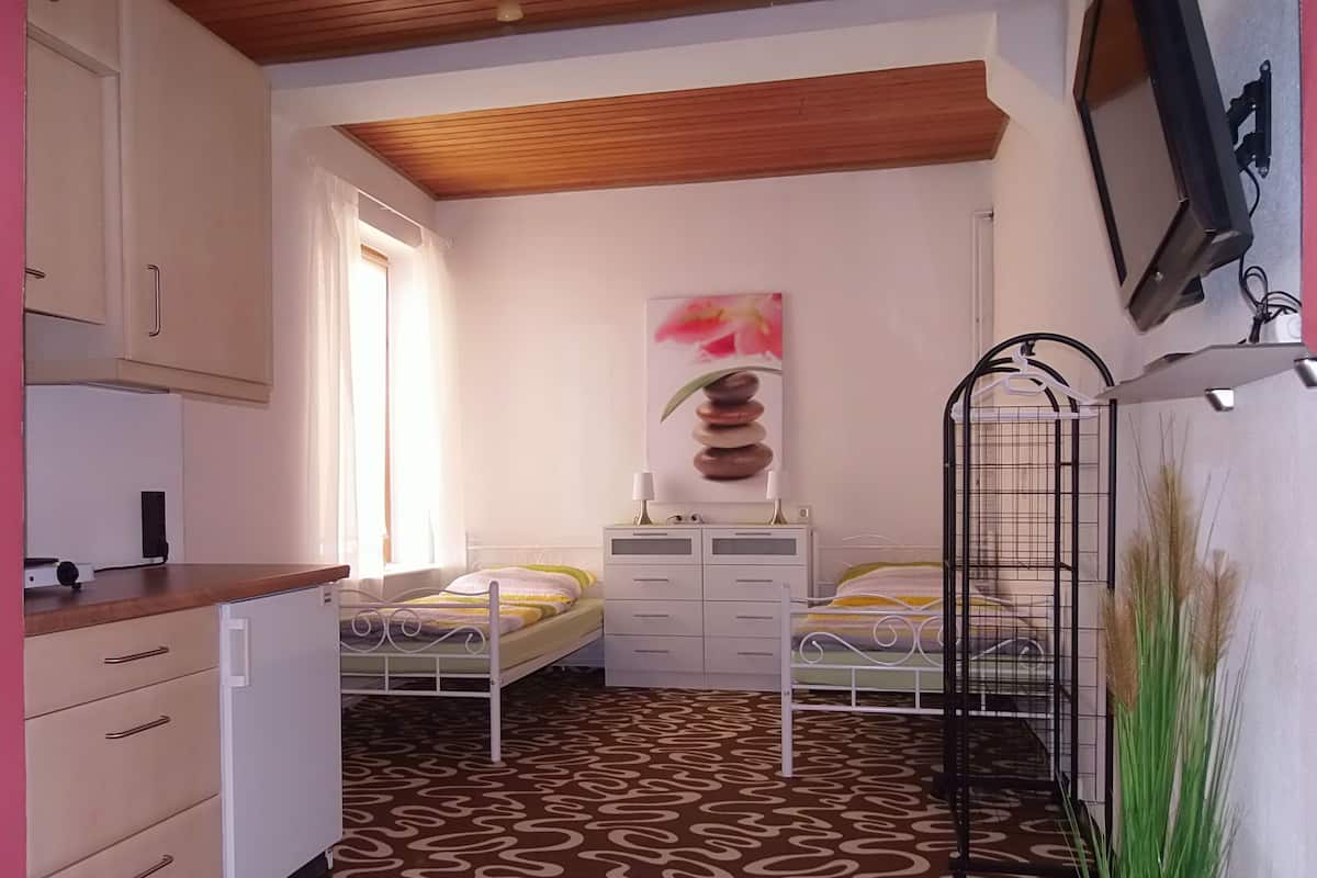 A small, well-lit bedroom with two single beds, a white dresser, a small desk, and a wall-mounted TV.