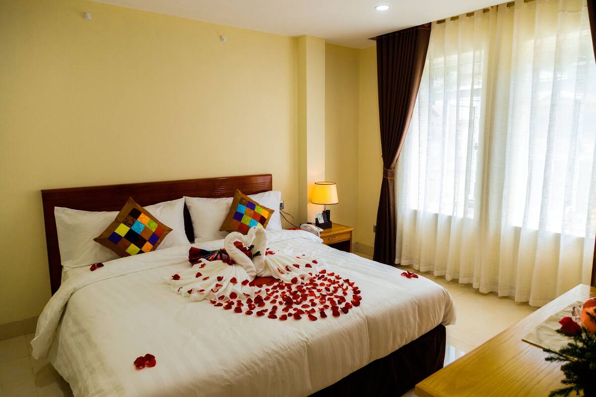 Una habitación de hotel con una cama grande, una mesita de noche con lámpara y un ventanal con cortinas.