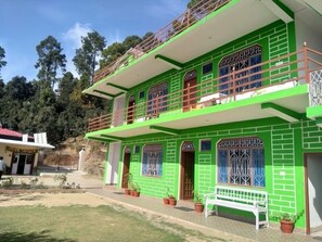 A two-story building with a green facade, multiple windows with decorative grills, and a balcony with a railing. - StayApart - Maa Anadmayee Guest House (Almora)