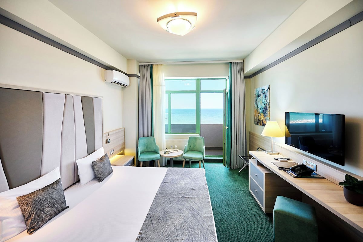 Ein Hotelzimmer mit einem großen Bett, einem Schreibtisch mit Fernseher, einem kleinen Tisch mit Stühlen und Meerblick durch ein Fenster.