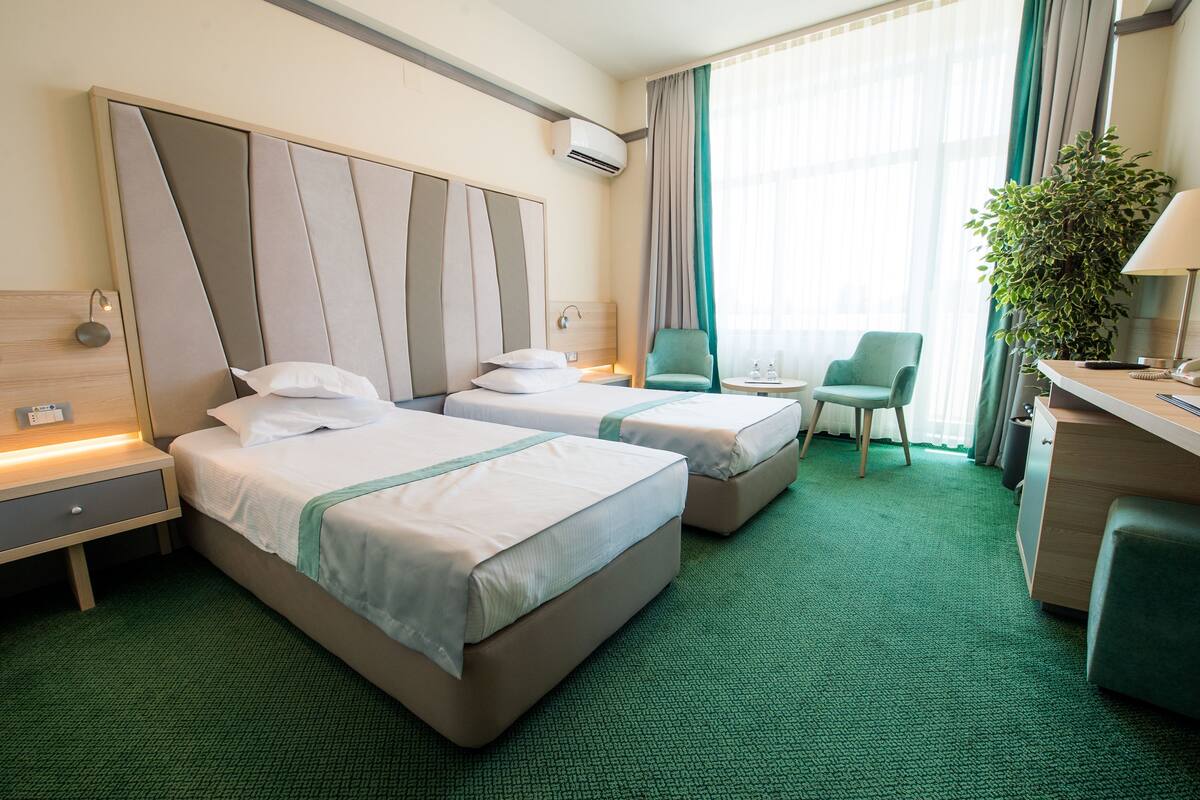 Ein Hotelzimmer mit zwei Betten, einem Schreibtisch, einem Sessel, einer Zimmerpflanze und einem Fenster mit Vorhängen.