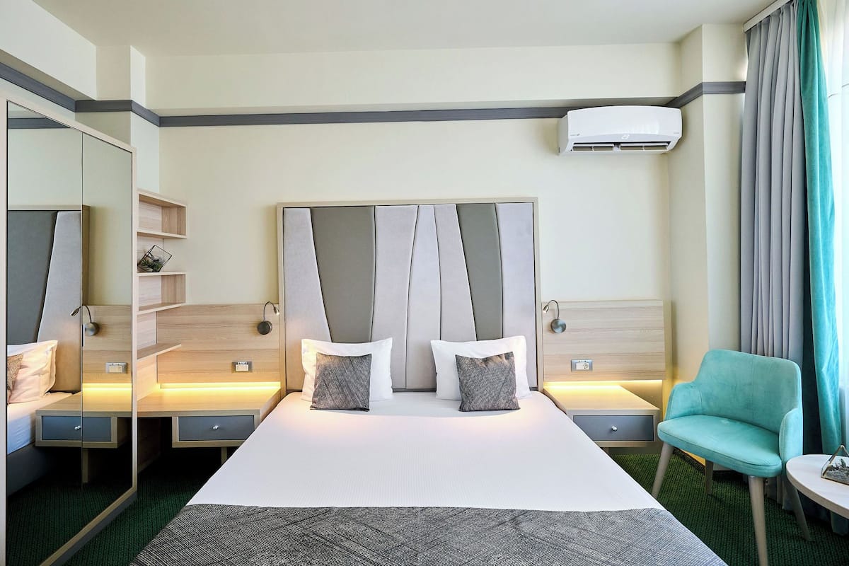 Ein Hotelzimmer mit einem großen Bett, Nachttischen, einem Sessel, einem Spiegel und einer Klimaanlage.