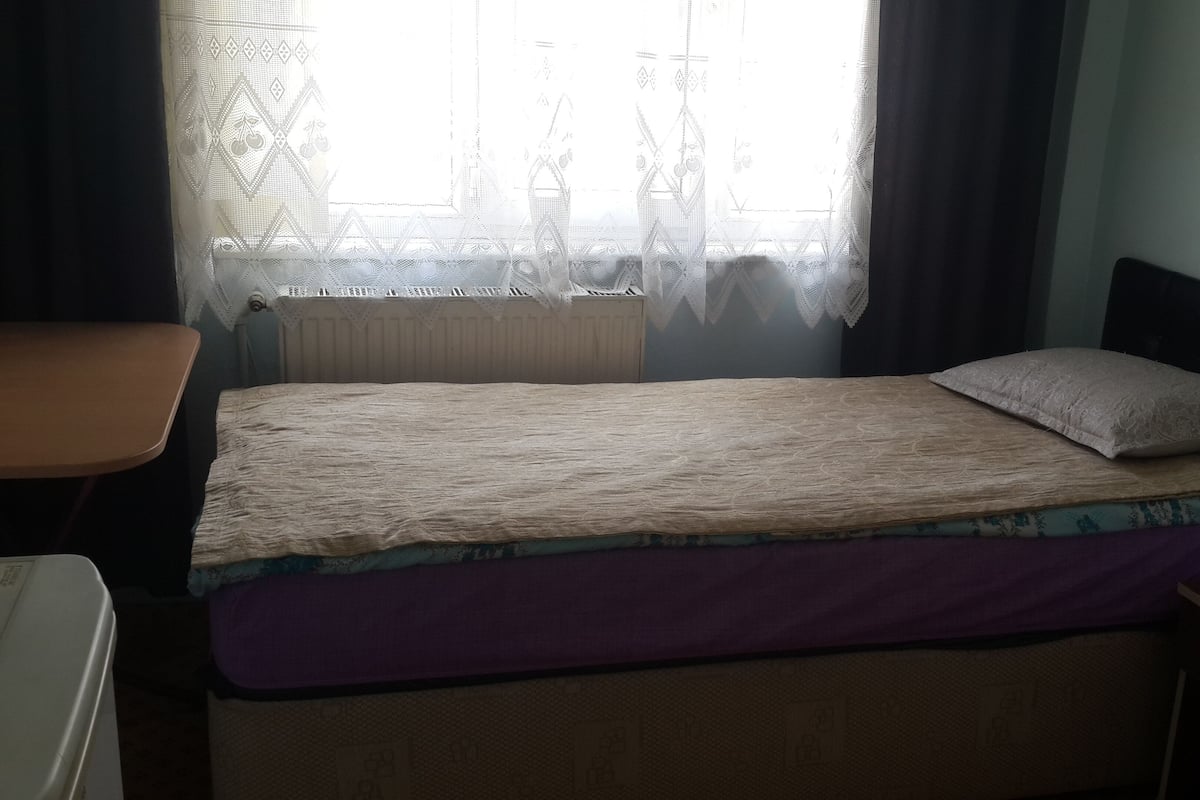 A single bed with a patterned bedspread, a pillow, and a headboard. There is a window with a lace curtain and a radiator below it. A small table is visible to the left of the bed.