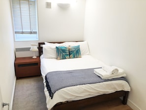 Een net opgemaakt bed met een bedsprei met een patroon, een bedlampje en een raam met zonwering.