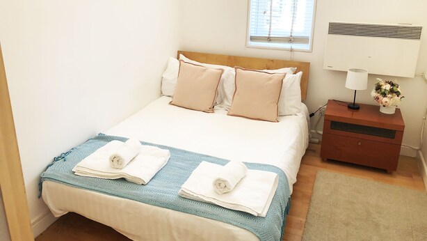 Un lit bien fait, recouvert d’une couvre-lit à rayures bleues et blanches, avec une tête de lit en bois et plusieurs oreillers. On y trouve une table de chevet sur laquelle sont posés une lampe et un vase de fleurs.
