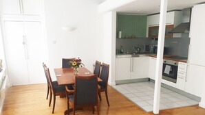 Un coin repas avec une table et des chaises en bois, un vase rempli de fleurs, et une cuisine avec des armoires blanches et un dosseret carrelé.