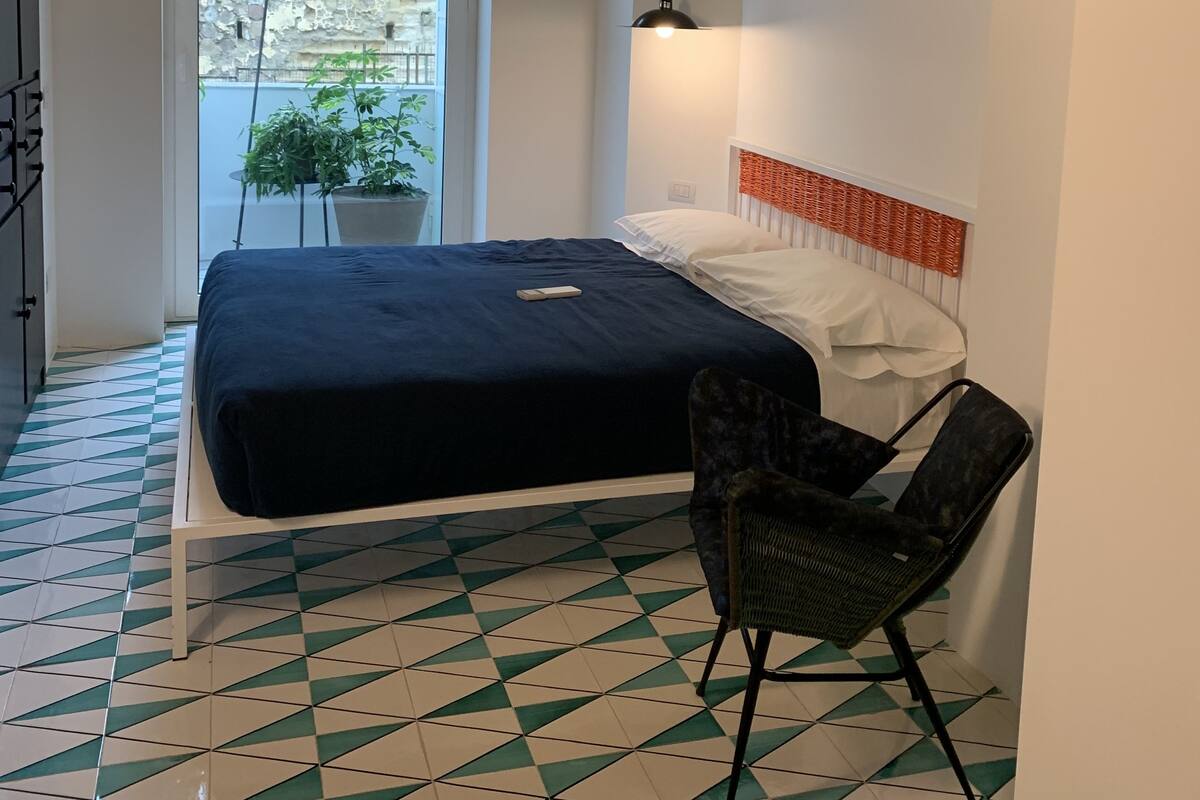 Quarto com cama, cadeira, luminária pendente, janela e vista para o verde lá fora.