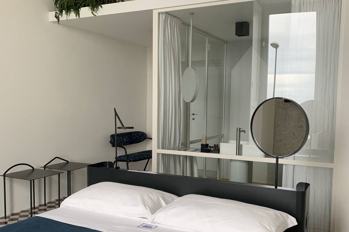 Quarto de hotel com cama, escrivaninha, cadeira, espelho e planta em vaso.