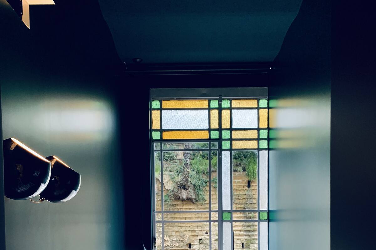 Quarto com cama, janela com vista para uma parede de tijolos e vegetação, e luminária fixada na parede.