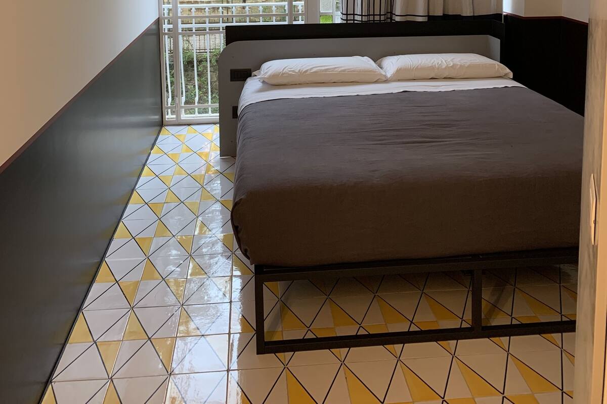 Quarto moderno com cama, luminária pendente, vista pela janela com cortinas e piso estampado.