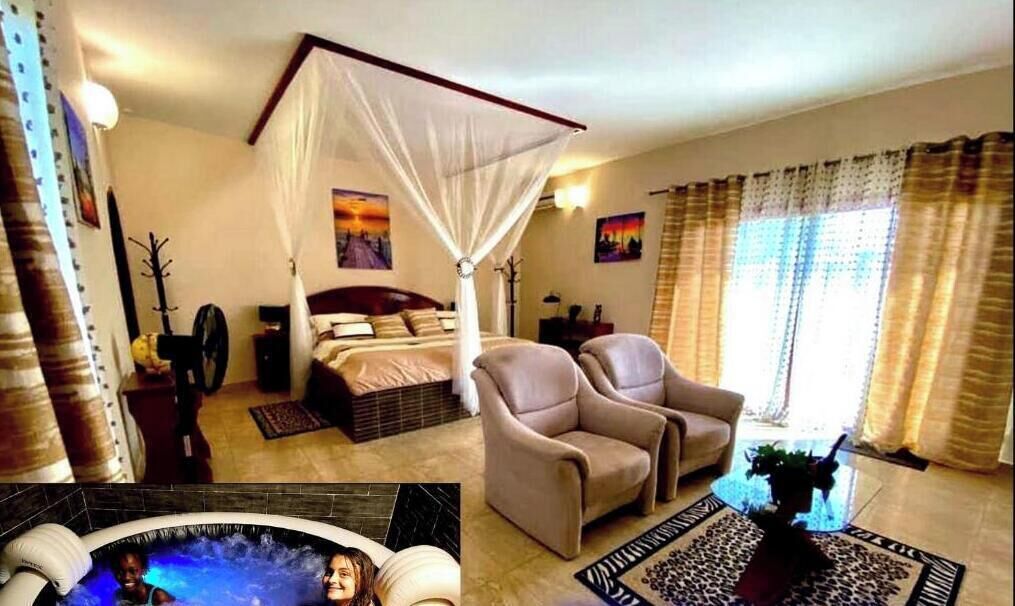 A bedroom with a four-poster bed, a sitting area with two chairs, a large window with curtains, and a hot tub in the corner.