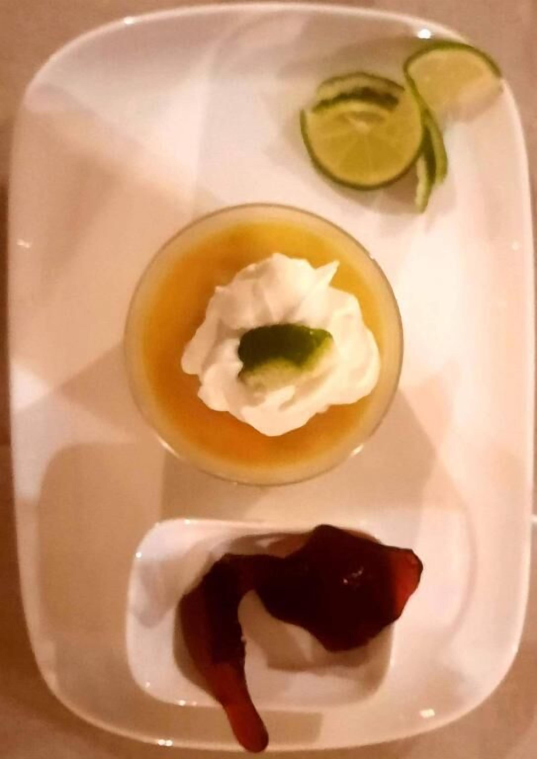 A dessert plate with a creamy dessert topped with a dollop of cream and a slice of lime, accompanied by a small dish of red sauce and lime slices.