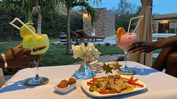 Un área de comedor al aire libre con una mesa preparada para dos personas, que incluye un plato de aperitivos, un vaso de bebida con una rodaja de limón y un jarrón decorativo.