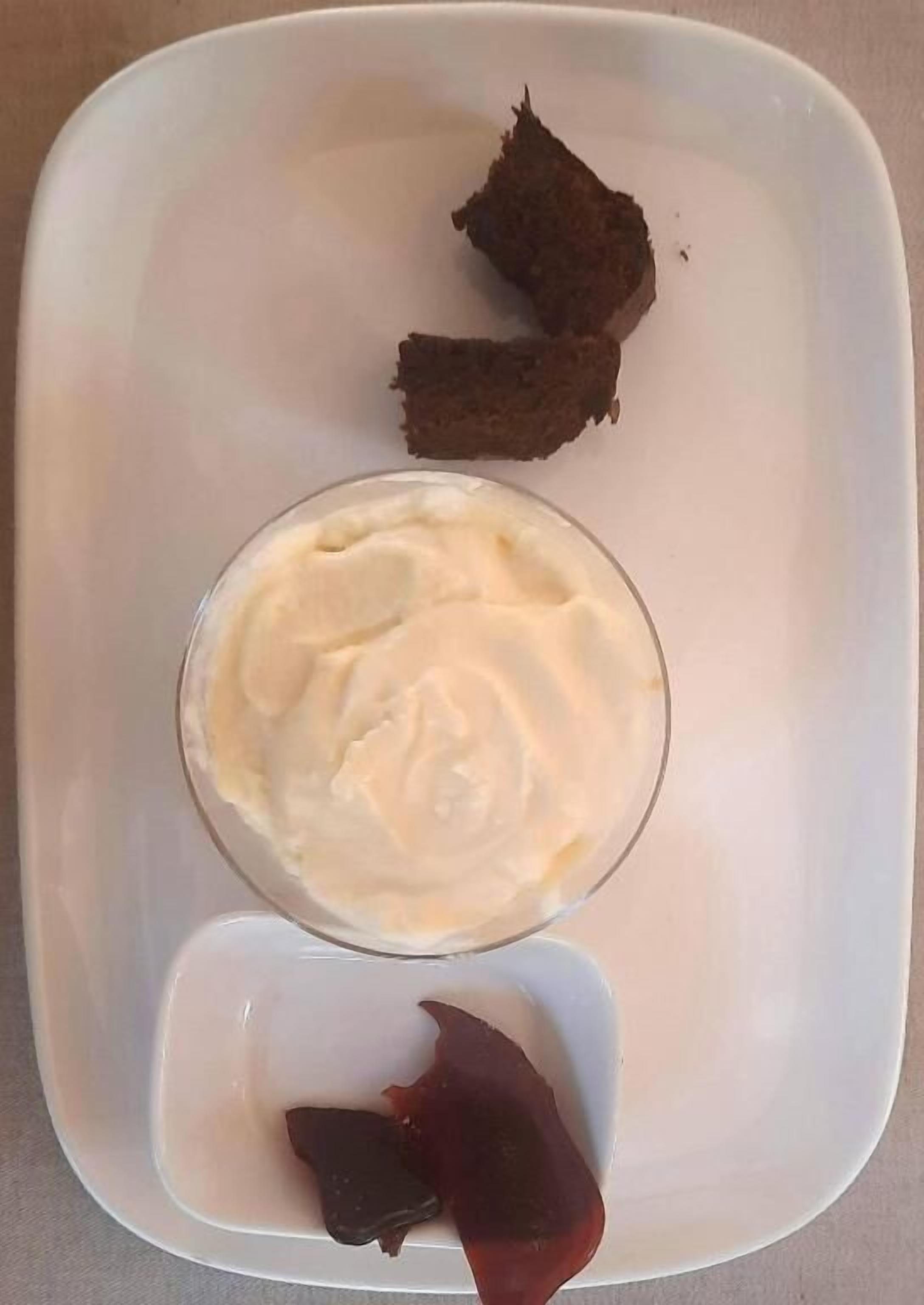 A white rectangular plate with a glass of white cream, two pieces of dark brown cake, and a small square dish with a dark red item.