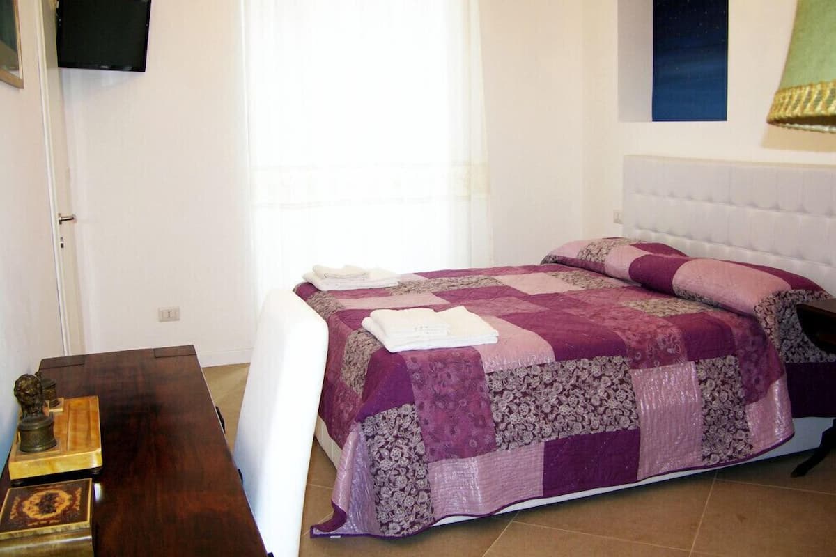 A bedroom with a bed, a nightstand, a flat-screen TV mounted on the wall, and a window with curtains.
