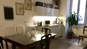 Ein Essbereich mit einem Glastisch, Holzsesseln, einer weißen Schrank mit Büchern, einer Kaffeemaschine und einer Zimmerpflanze.