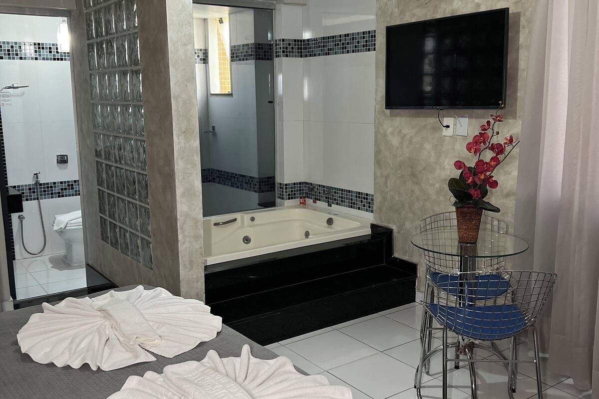 Een moderne hotelkamer met een groot bed, een douchecabine met glazen wanden, een badkuip, een aan de muur bevestigde televisie en een klein tafeltje met stoelen.