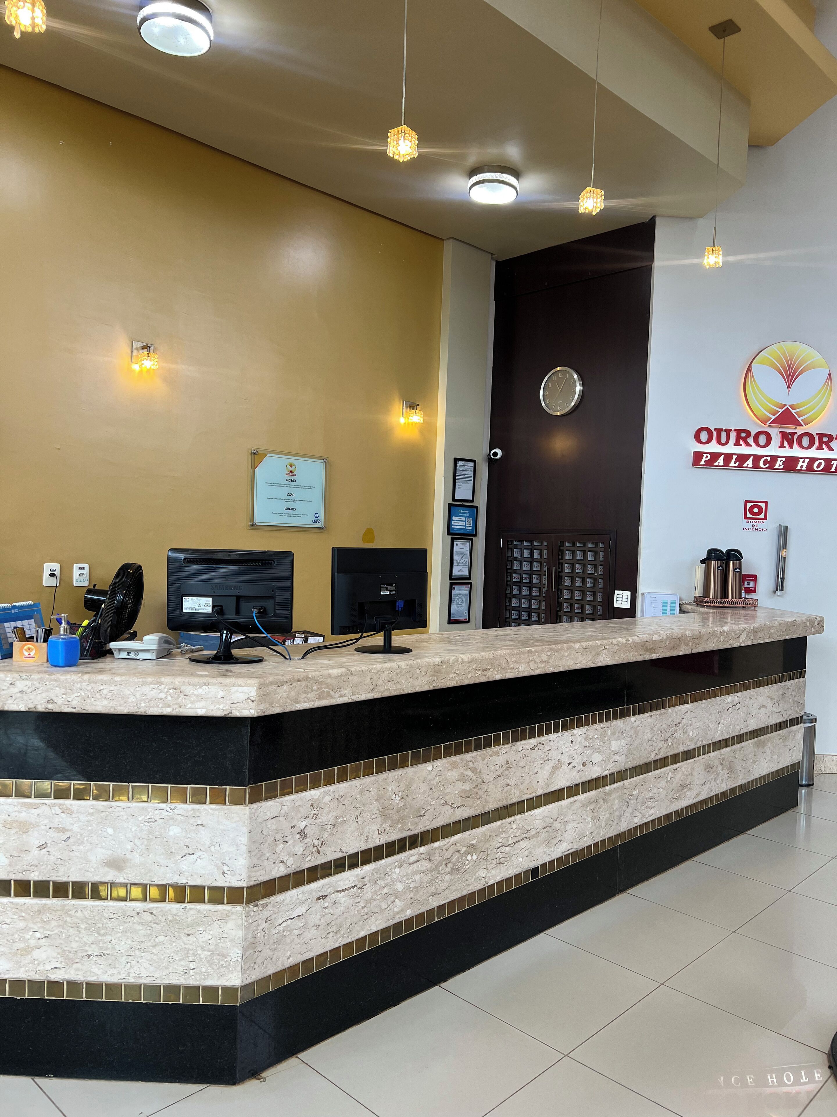 A hotel reception desk with a marble counter, a clock, and a sign for Ouro Norte Palace Hotel.