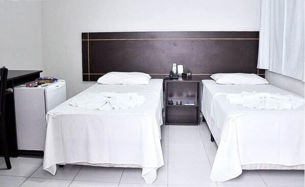 Quarto de hotel com duas camas de solteiro, uma pequena mesa de cabeceira com abajur e uma escrivaninha com cadeira.