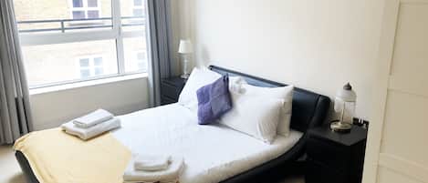 A hotel room with a bed, a nightstand, a lamp, and a view of the outside.