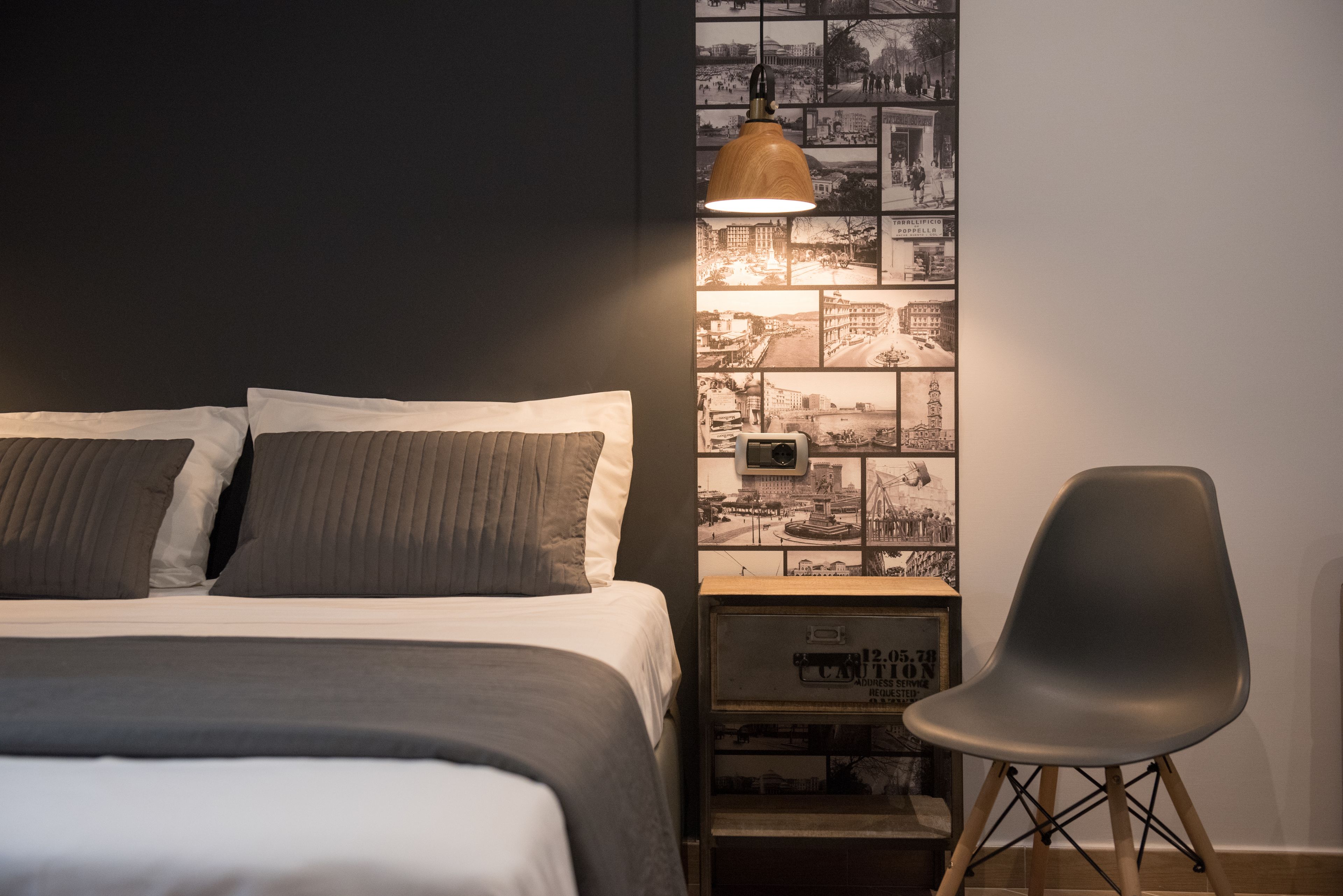 A neatly made bed with two pillows, a modern lamp, a chair, and a wall with a collage of black and white photos.