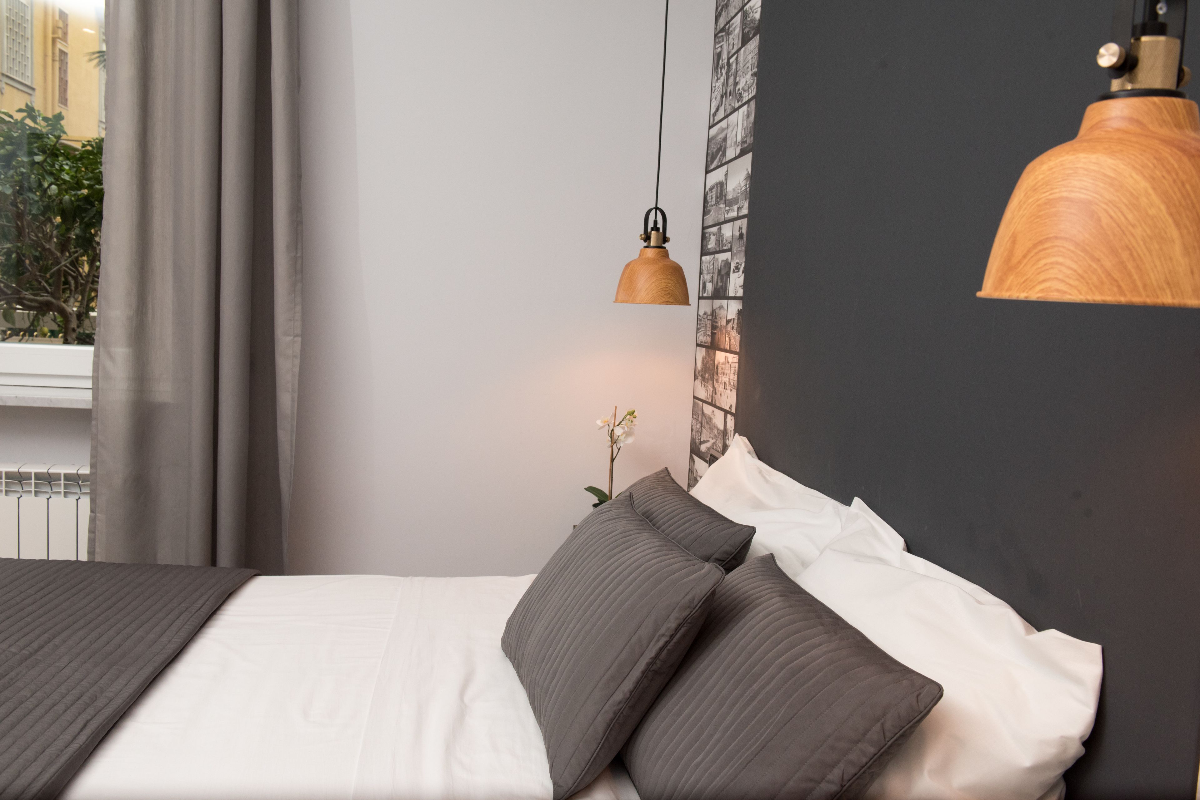 A neatly made bed with dark and light pillows, a hanging pendant light, and a wall with a black and white photo collage.