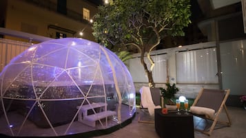 A transparent geodesic dome structure with seating inside, surrounded by a garden with a tree and a small table with drinks.