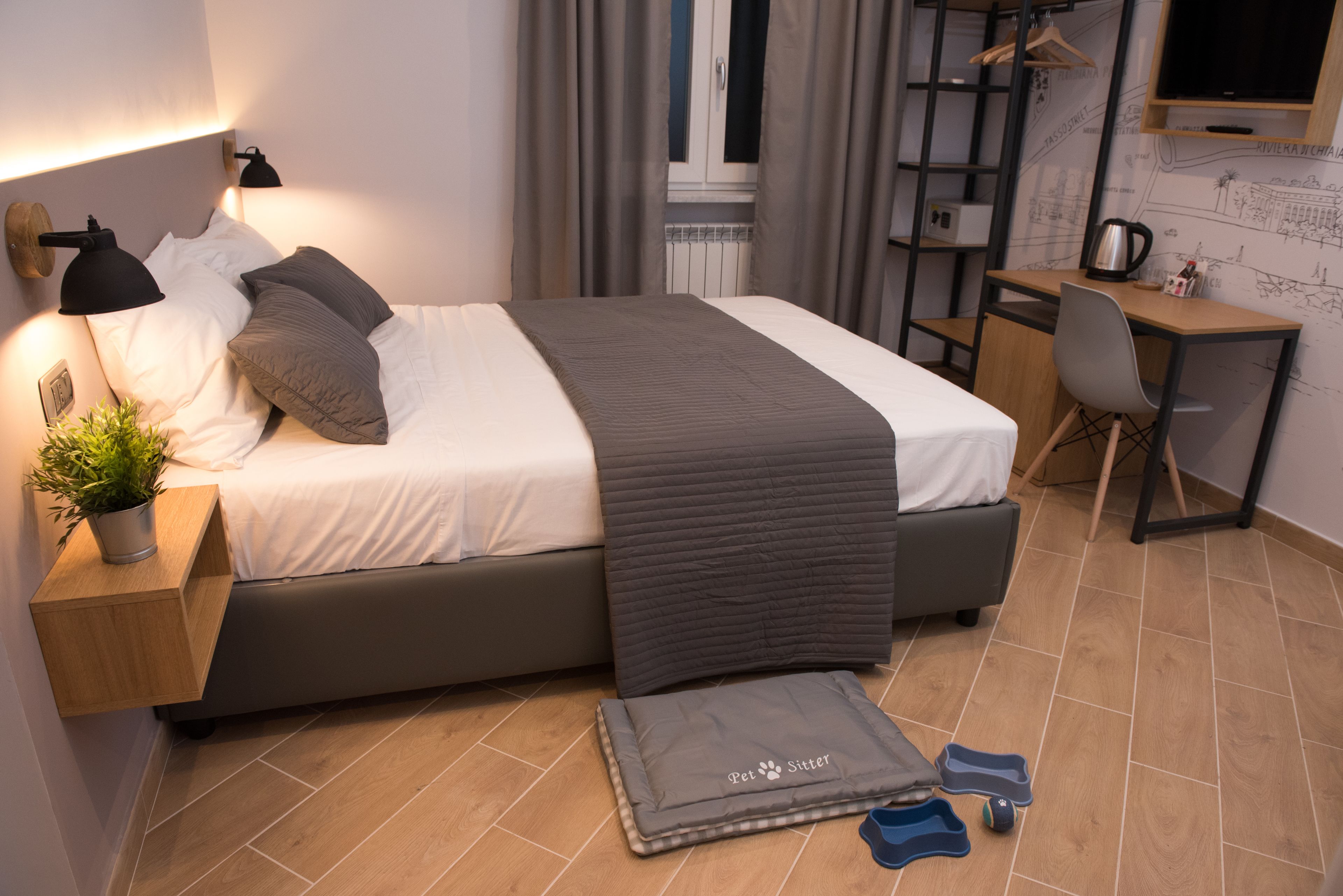 Habitación de hotel moderna con cama, mesita de noche, lámpara, planta en maceta, cama para mascotas, pantuflas y un escritorio con silla.