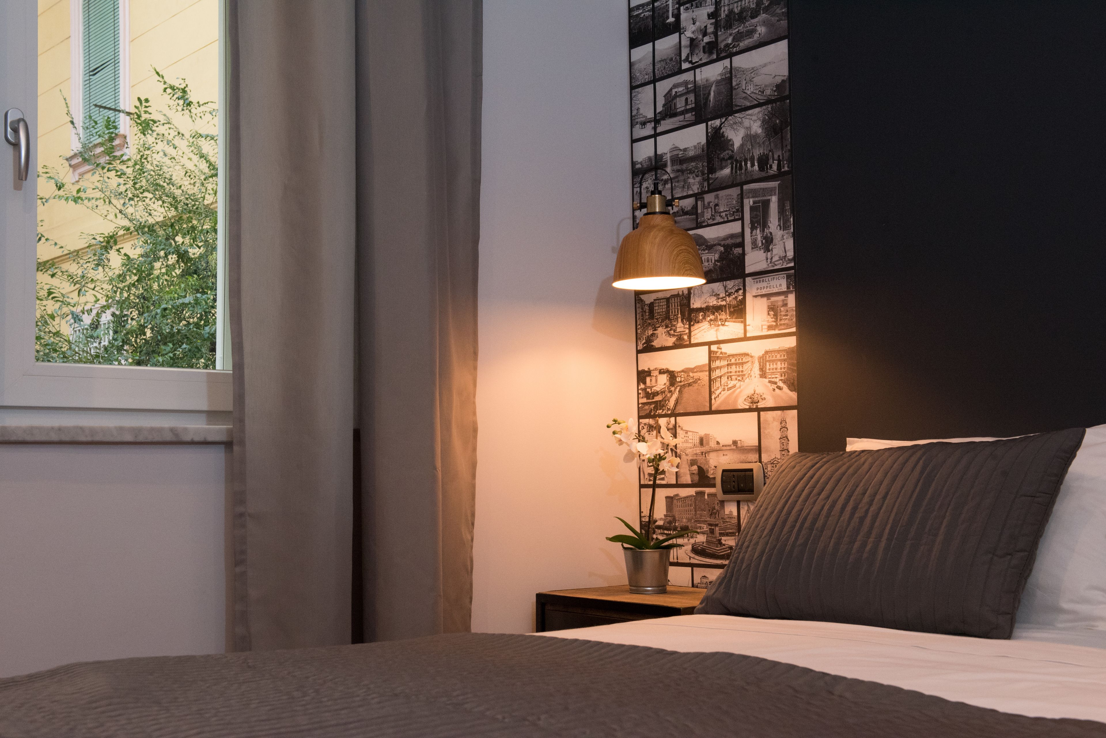 Quarto com cama, mesa de cabeceira, luminária, parede com fotos em preto e branco e janela com cortinas.