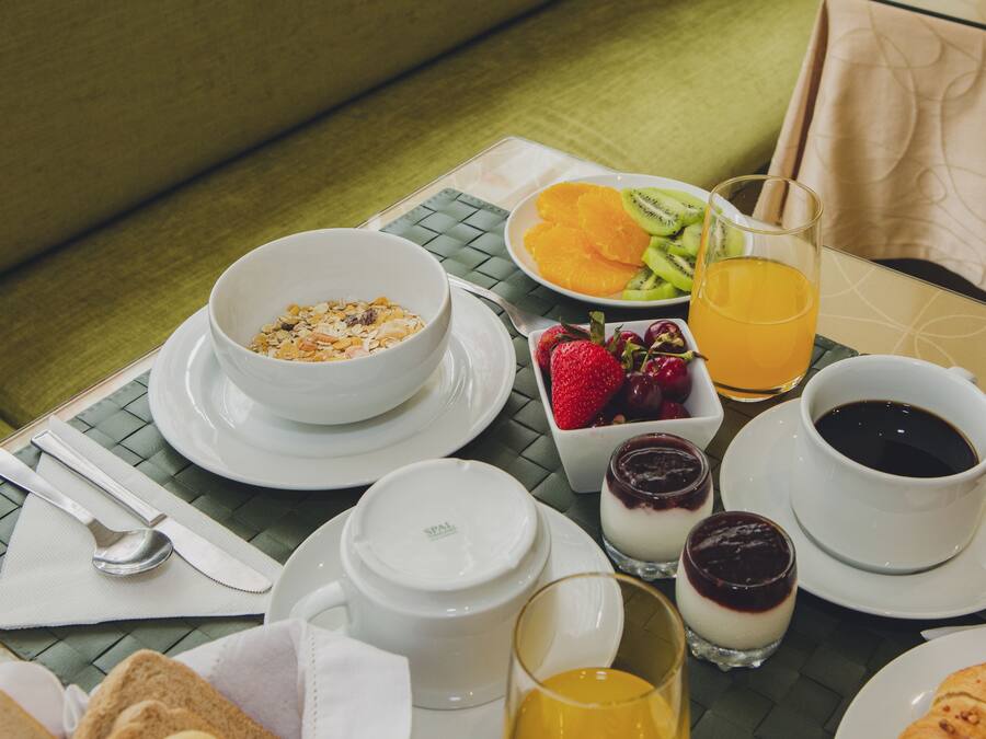 A breakfast setting with a bowl of cereal, orange slices, kiwi, strawberries, a glass of orange juice, a cup of coffee, and a dessert on a table.