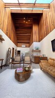 A spacious living area with a high ceiling, wooden paneling, a sofa, chairs, a coffee table, and a kitchen area with a sink and countertop.
