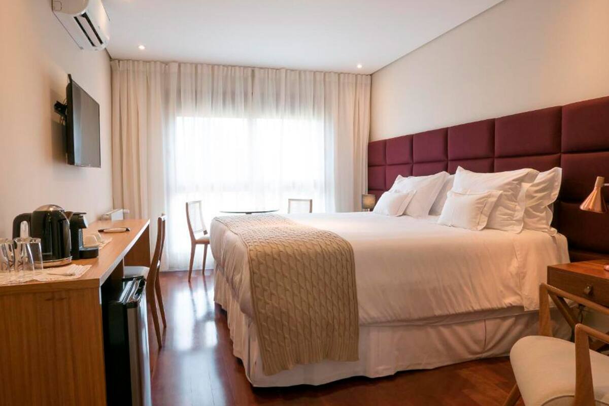 A hotel room with a bed, a desk with a kettle, a chair, a TV mounted on the wall, and a window with curtains.