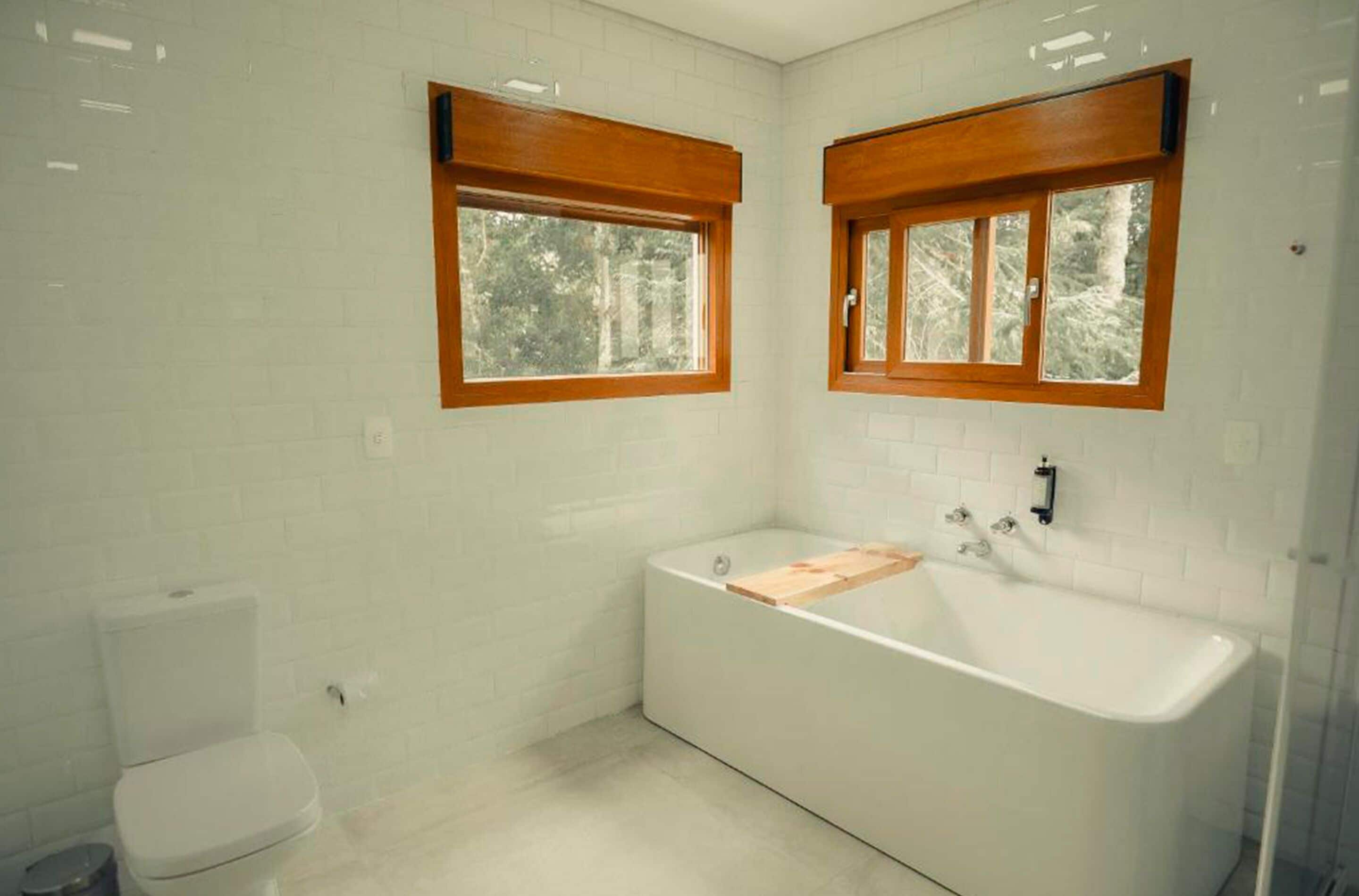 A bathroom with a white bathtub, a toilet, and two windows with wooden frames.