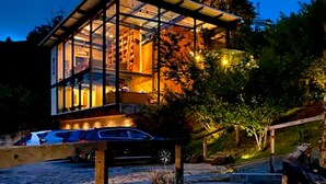 A modern glass house with a wooden deck, surrounded by trees and a car parked nearby. - Hotel Boutique Quebra-Noz Conforto e Natureza (Campos do Jordão)