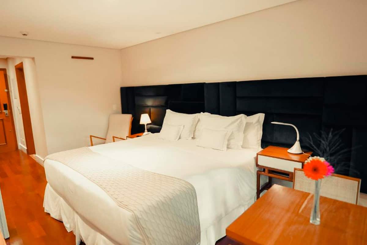 A hotel room with a large bed, a bedside table with a lamp, a chair, and a small table with a vase.
