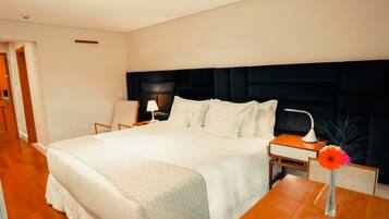 Quarto de hotel com uma cama grande, uma mesa de cabeceira com luminária, uma cadeira e uma pequena mesa com um vaso.