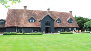 A large, dark-colored building with a steeply pitched roof and multiple chimneys. It features a well-maintained lawn in front and a patio area with outdoor furniture. - The Tithe Barn (Maidenhead)