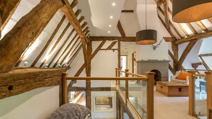 A loft bedroom with exposed wooden beams, a glass staircase, a fireplace, and a cozy seating area. - The Tithe Barn (Maidenhead)