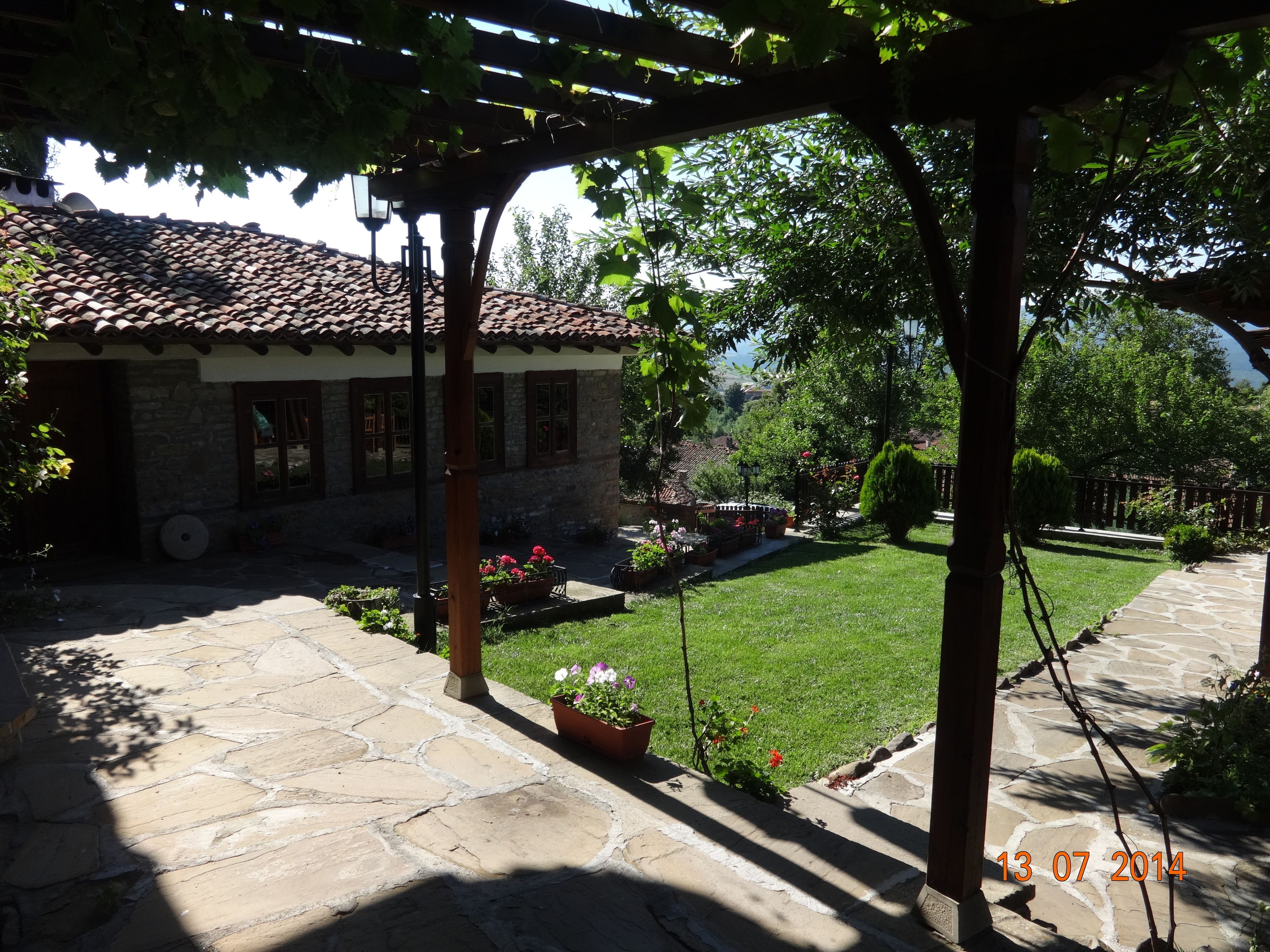 Un patio empedrado con una pérgola, una casa con techo de tejas y un césped cuidado.