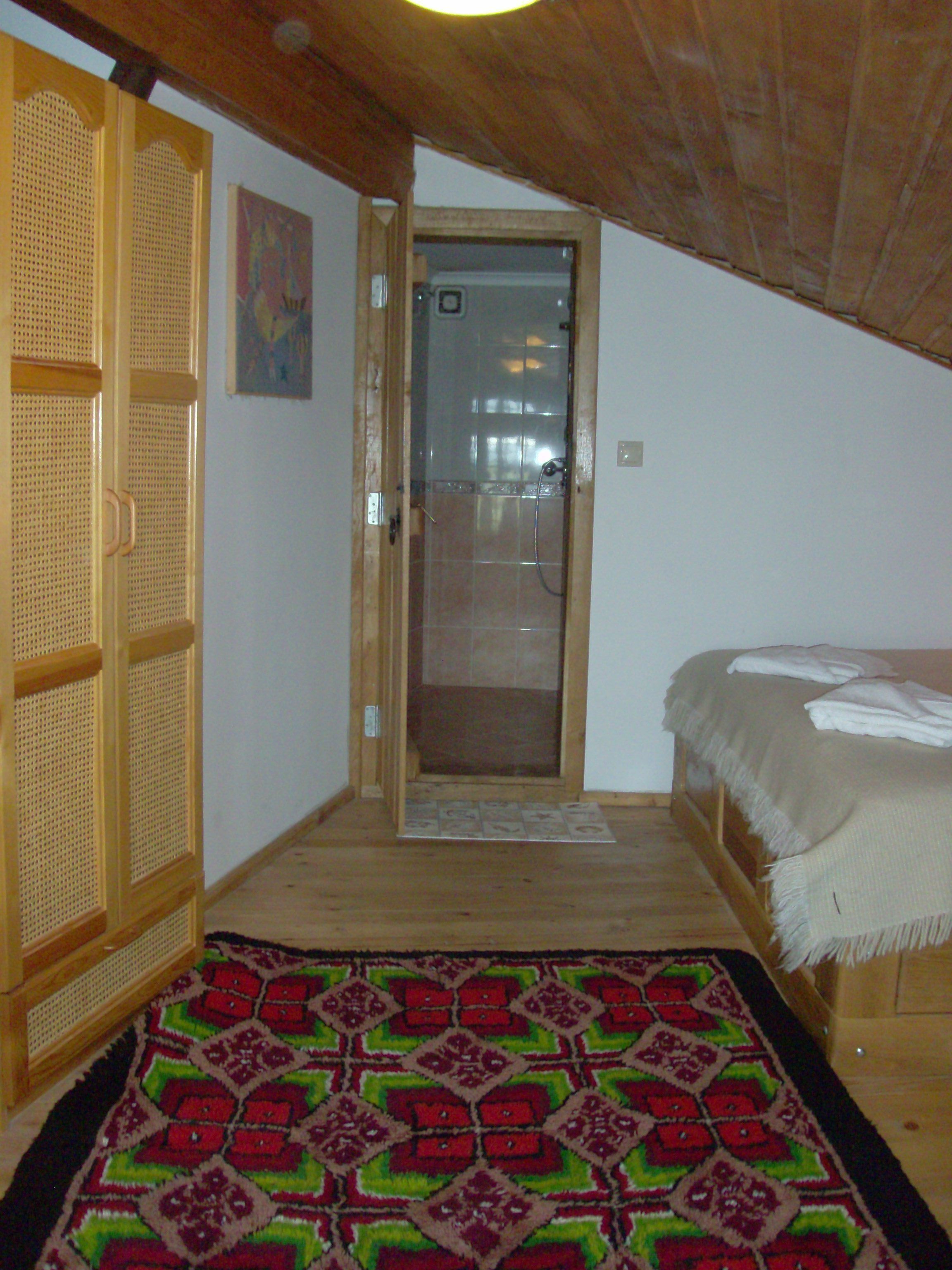 Une chambre avec un plafond en bois, un lit recouvert d’une couverture blanche, un tapis à motifs et une armoire en bois.