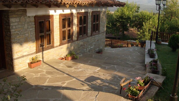 A stone building with a tiled roof, multiple windows, and a courtyard with potted plants and a bench. - Zarkovata Kashta (Kotel)