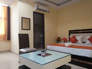 Une chambre d’hôtel comprenant un lit, une table basse en verre, une chaise, une porte, un appareil de climatisation et une petite table sur laquelle est posé un vase de fleurs.