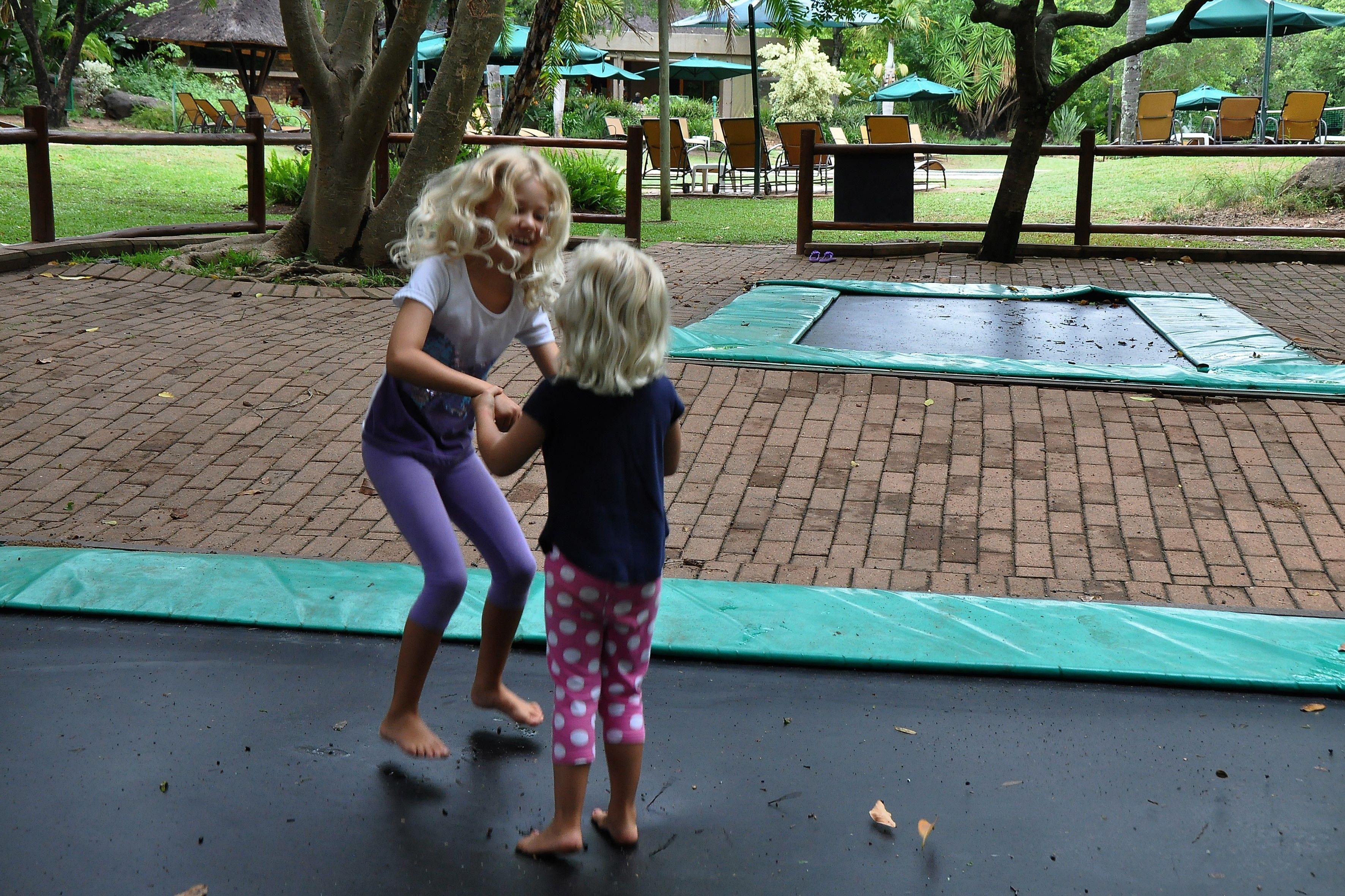 Dos niños jugando en un trampolín en un espacio exterior con un camino de ladrillos, rodeado de follaje y mobiliario de exterior.
