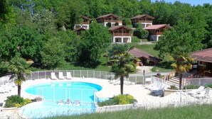 A resort with a swimming pool surrounded by lounge chairs, set against a backdrop of forested hills and multiple chalet-style buildings. - Village du Soleil (Mauroux)
