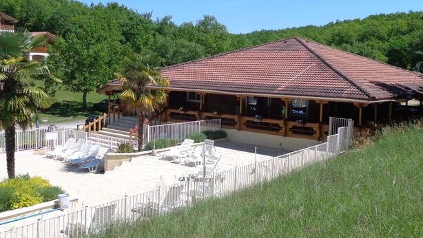 A building with a tiled roof, a covered patio, and a fenced outdoor area with lounge chairs. - Village du Soleil (Mauroux)