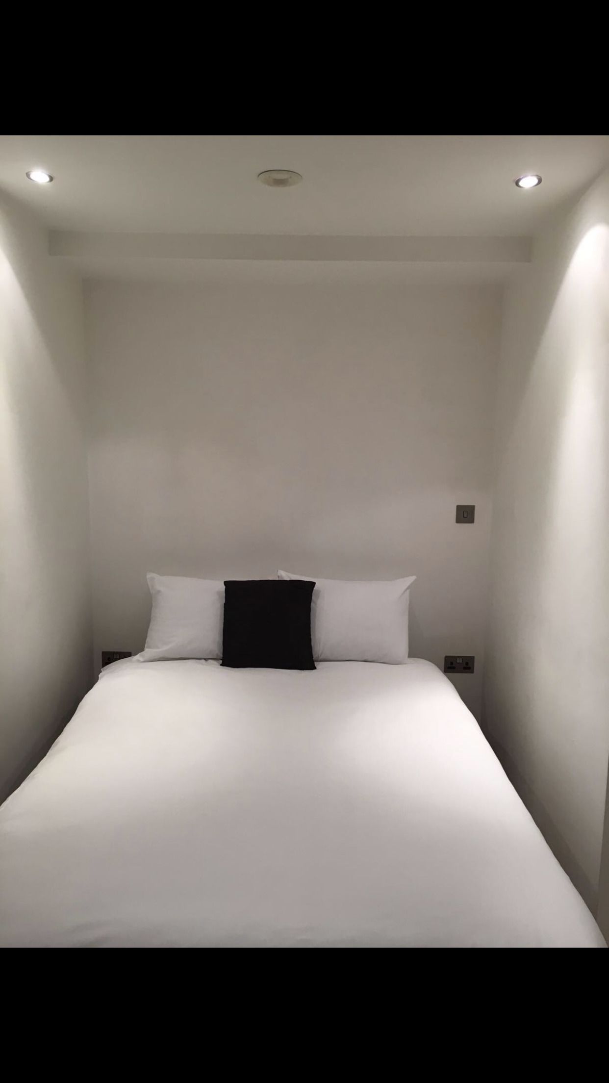 A small, narrow hotel room with a single bed, white walls, and a ceiling light.