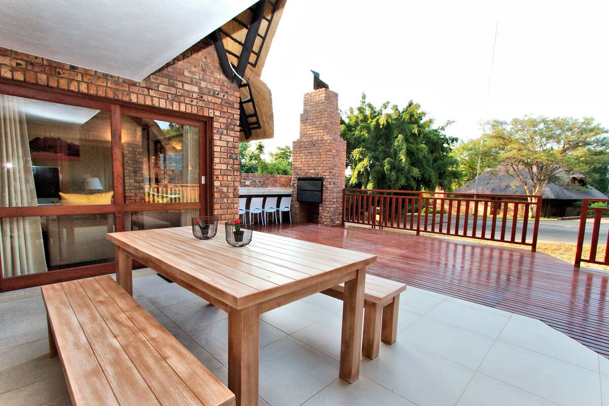 An outdoor patio with a wooden table and benches, a brick fireplace, and a view of the surrounding landscape.