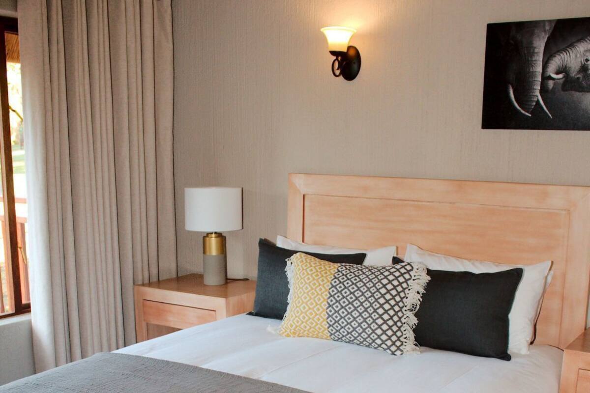 A neatly made bed with a headboard, a bedside lamp, a wall-mounted air conditioner, and a painting on the wall.