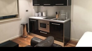 A compact kitchen with modern appliances, a dining area with a black leather chair, and a small trash bin. - The Greengate Apartment (Salford)