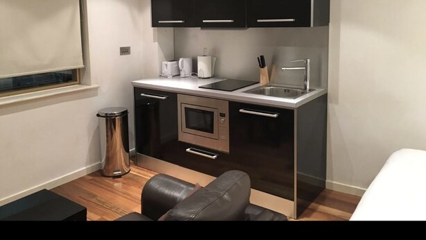 A compact kitchen with modern appliances, a dining area with a black leather chair, and a small trash bin. - The Greengate Apartment (Salford)